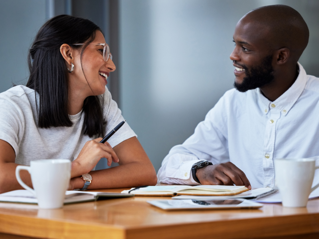 a female real estate agent with medium skin and glasses discusses the loan officer and real estate agent relationship with a dark skinned male loan officer at a table over coffee