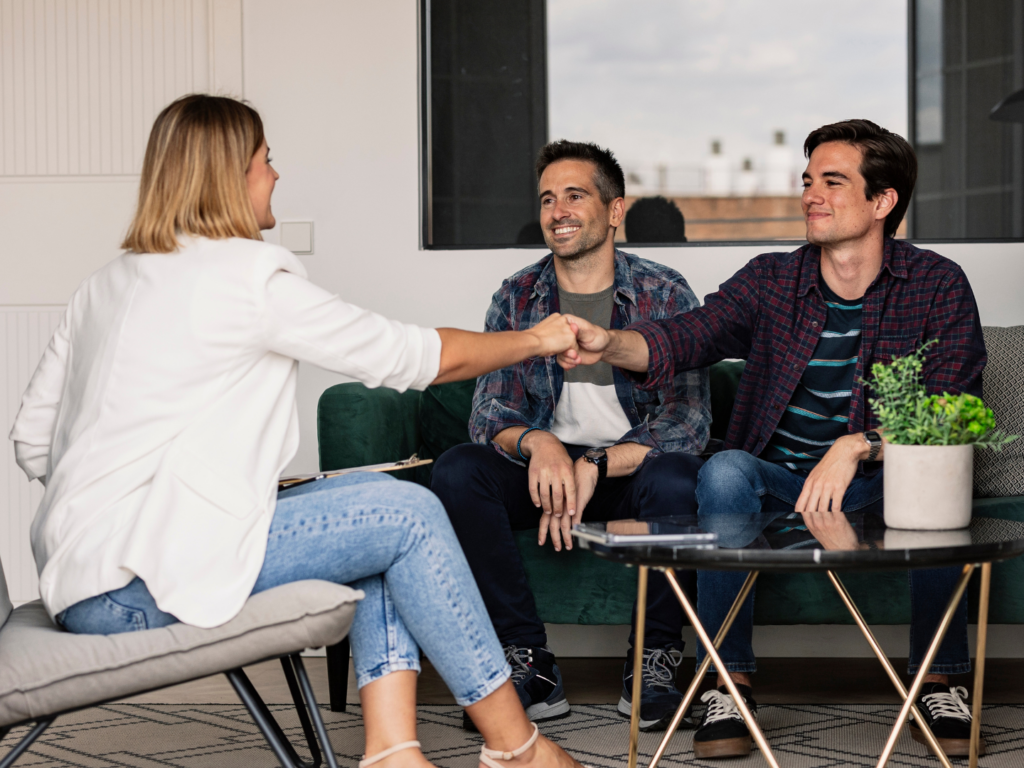 a same sex male couple, both with medium-tanned skin and dark hair, meet with a female real estate agent who specializes in working with first-time homebuyers