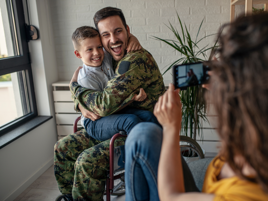 military father in army fatigues and a wheel chair holds his son in his lap as woman takes a picture of them moving in to a new home purchased with a veterans affairs loan